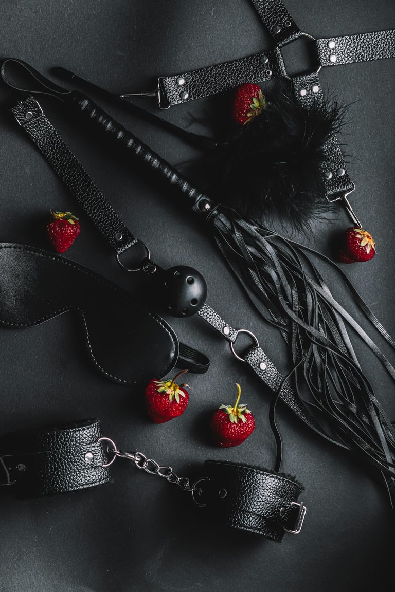 Flat lay of black leather BDSM accessories with metal studs and red strawberries on dark gray background