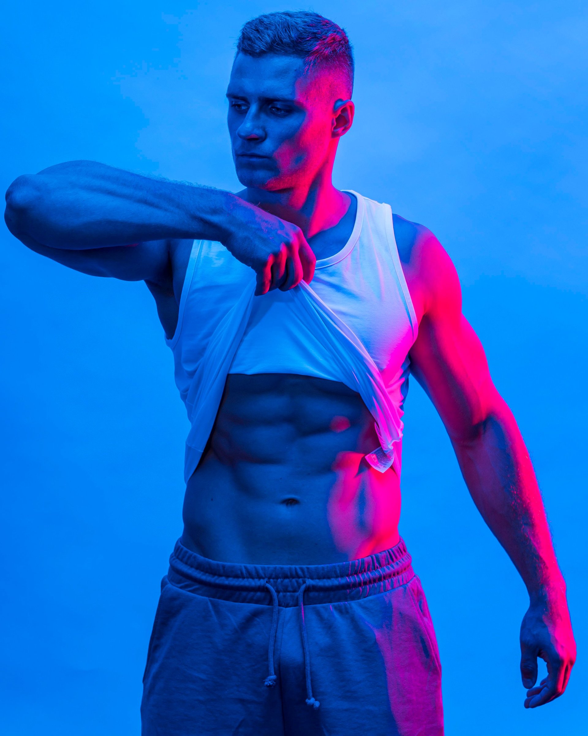Athletic man in white tank top posing against blue and pink neon background