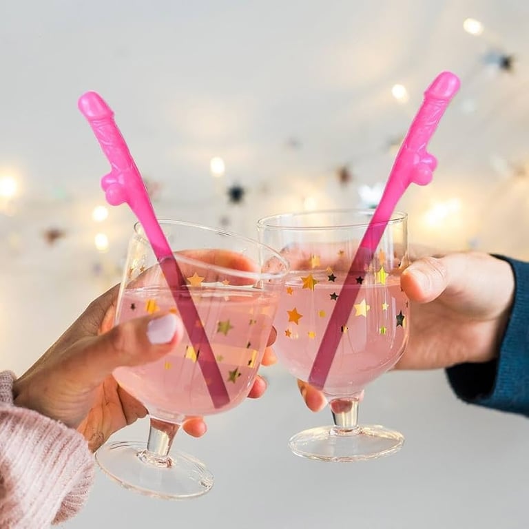 Two hands holding festive pink cocktails with novelty straws against a bokeh background with confetti and party lights